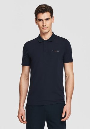 Mężczyzna noszący dopasowaną czarną koszulkę polo z krótkim rękawem, kołnierzykiem i małym białym logo na lewej piersi, stojący na jasnym jednolitym tle.