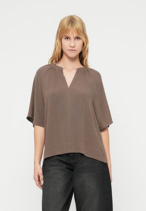 Braunes kurzärmliges Oberteil mit V-Ausschnitt und leichter Faltenlegung an den Schultern, aus einem strukturierten Material, kombiniert mit dunklen Jeans.