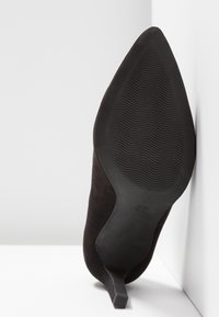 Chaussure à talon haut noire à bout pointu, inclinée vers le haut, montrant une semelle en caoutchouc texturée avec la taille 37 embossée près du talon.