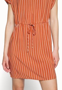Rostfärgad klänning med vita vertikala ränder, korta ärmar, rynkad midja och knytdetalj. Tillverkad av ett mjukt, lätt tyg.