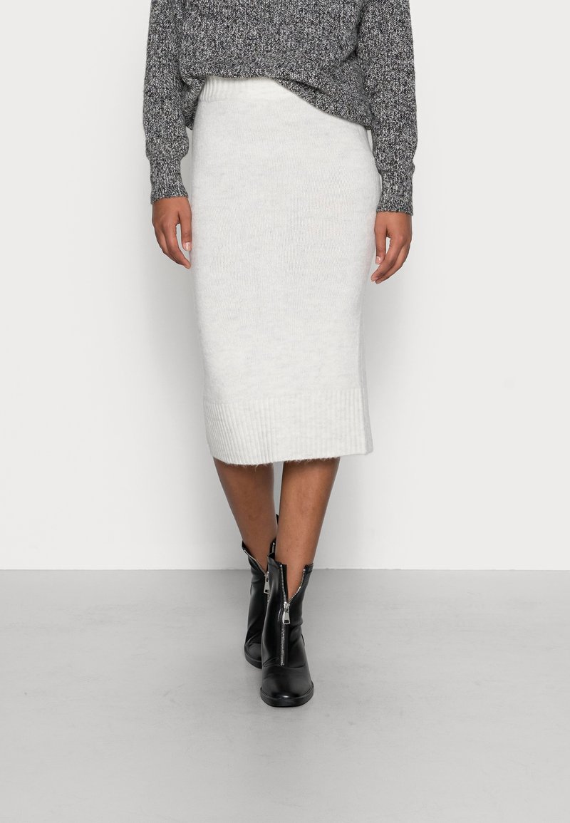 Personne portant une jupe midi en tricot blanc, un pull gris chiné et des bottines noires à fermeture éclair frontale, debout devant un fond uni.