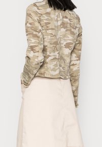Camouflage jacket in tan and green tones, featuring a cropped design and frayed hem. Worn over a light beige skirt.
