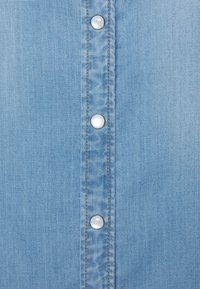 Camicia in denim blu chiaro con una fila di bottoni a pressione argentati lungo il centro e cuciture a contrasto color sabbia, caratterizzata da una texture liscia.