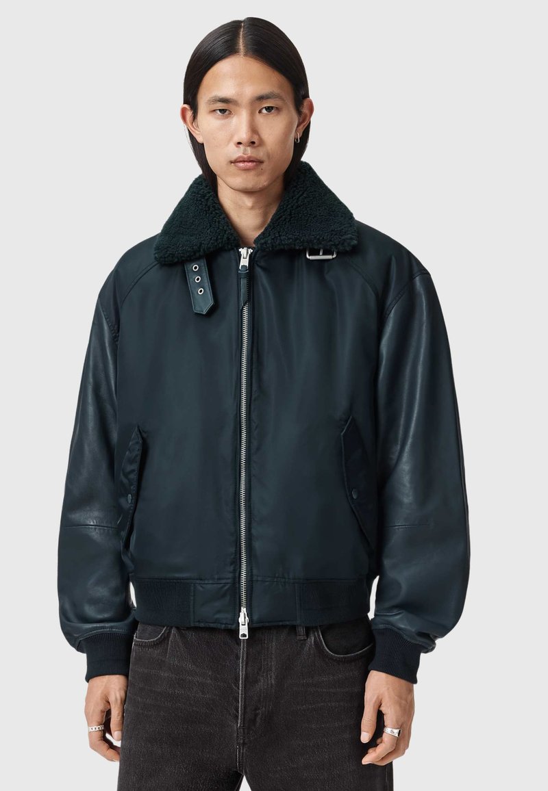 Tummansininen bomber-takki, jossa suuri, teksturoitu musta tekoturkiskaulus; takissa on vetoketjusuljenta, sivutaskut sekä resorit hihansuissa ja helmassa.