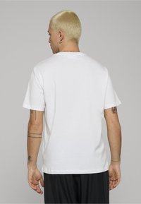 Camiseta blanca de algodón con un corte estándar, mangas cortas y cuello redondo, vista desde atrás. Presenta tatuajes visibles.