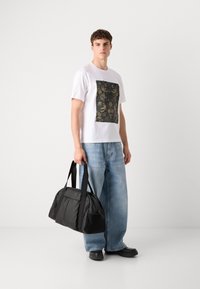 Weißes T-Shirt mit einem schwarzen Blumenaufnäher, lockere hellblaue Jeans und eine schwarze Reisetasche mit eingeprägtem "BOSS". Schwarze Schuhe.