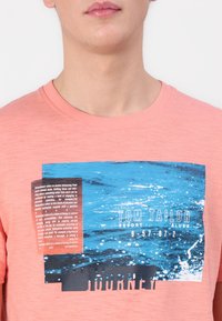 Koral t-skjorte med et rektangulært grafisk trykk som viser blå og svarte havmotiver, "TOM TAILOR"-tekst og et strukturert merke på siden.