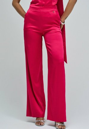Mujer con pantalones anchos de color rosa brillante con bolsillos, combinados con sandalias plateadas de tacón alto y tiras, de pie sobre un fondo sencillo.
