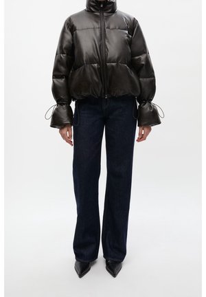 Osoba w czarnej skórzanej kurtce puchowej ze ściągaczami na rękawach, ciemnych prostych jeansach i spiczastych czarnych butach, stojąca na białym tle.