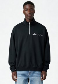 Sweatshirt noir à demi-fermeture éclair avec "Amour" en cursive blanche sur la poitrine. Comprend des poignets et un bas côtelés, fabriqué en coton doux.