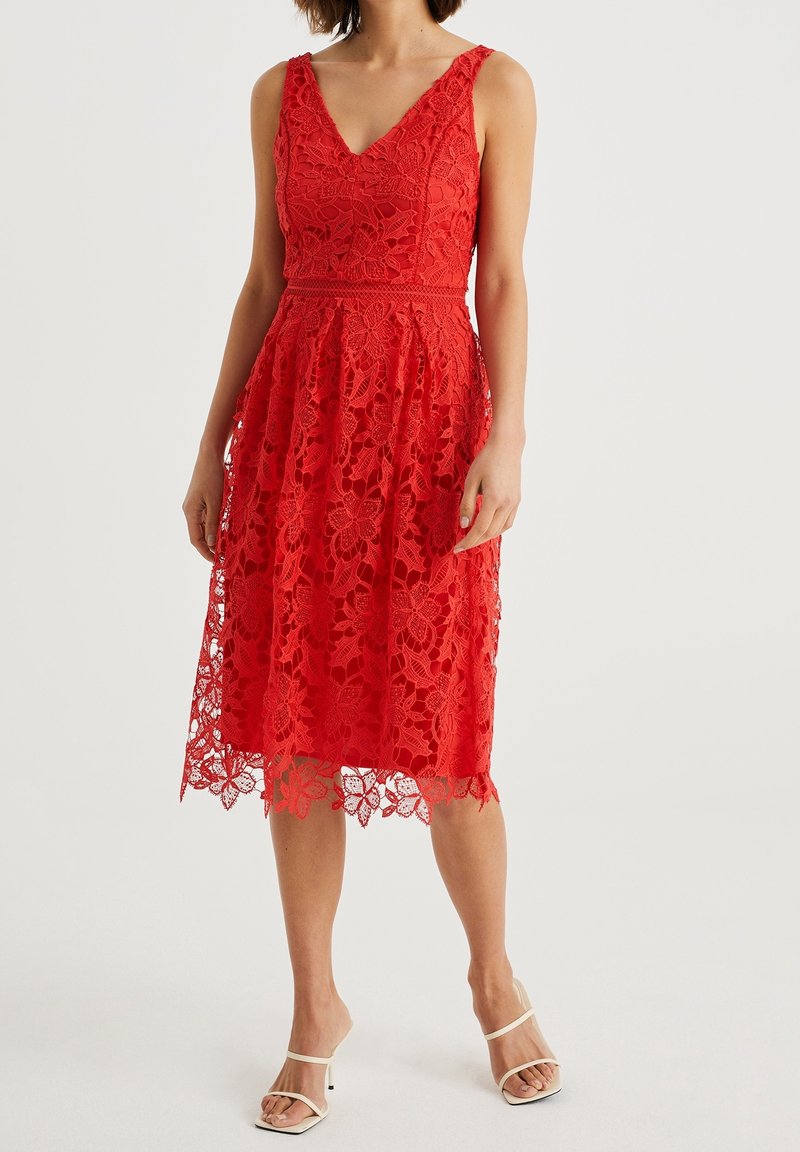 Robe rouge en dentelle avec un col en V, design sans manches, motif floral et ourlet festonné, associée à des sandales blanches à brides.