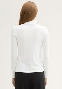 Weißes Langarm-Turtleneck-Oberteil mit glatter Textur, figurschmeichelndem Design und subtilen Rippenmustern, kombiniert mit schwarzen Hosen.