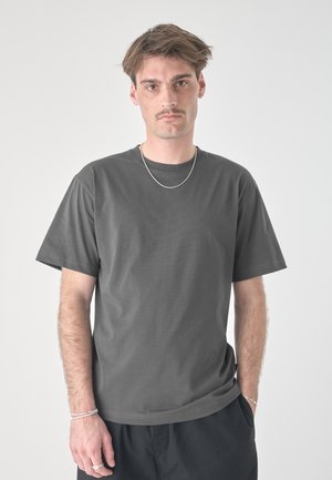 Jonge man met kort bruin haar en snor, die een grijs T-shirt, zilveren ketting, armbanden en ring draagt, staand tegen een effen lichte achtergrond.