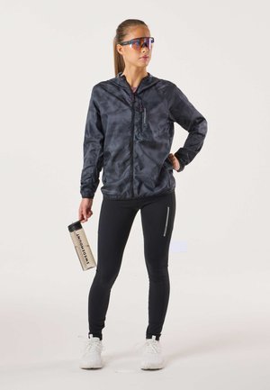 Ľahká čierna bunda so vzorom a predným zipsom, v kombinácii s čiernymi legínami. Drží priehľadnú fľašu na vodu s nápisom. Biele športové topánky.