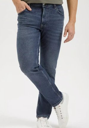 Mężczyzna w ciemnoniebieskich dżinsach z jedną ręką w kieszeni i białych trampek, stojący ze skrzyżowanymi nogami na jednolitym tle.