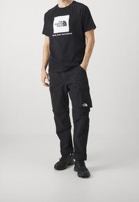 Osoba v černém tričku The North Face a cargo kalhotách, s turistickými botami, stojí s jednou rukou v kapse na bílém pozadí.