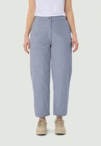 Osoba u svijetloplavim ravnim hlačama i bež tenisicama na vezanje, stoji s opuštenim rukama uz tijelo ispred jednostavne pozadine.