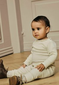 Beige, struktureret strikoutfit med ribbet mønster og volumiøse detaljer. Brune læderstøvler med snørebånd. Barn siddende på trægulv.