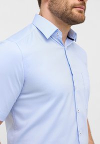 Korte mouwen, knoopsluiting shirt van lichtblauwe stof met een subtiele textuur, voorzien van een kraag, borstzak en witte knopen.