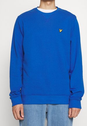 Hombre vistiendo un sudadera azul brillante de cuello redondo con un logo de pájaro amarillo en el pecho y jeans azul claro, contra un fondo blanco.