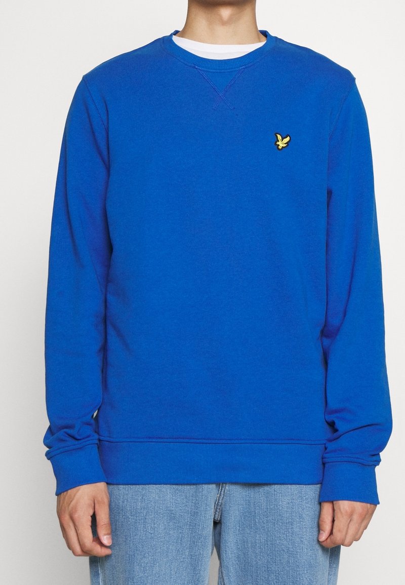 Man med ljusblå crewneck-tröja med gult fågel-logotyp på bröstet och ljusblå jeans mot vit bakgrund.