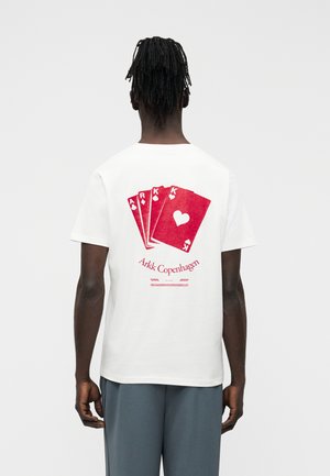 Άνδρας με πλεγμένα μαλλιά κοιτάζει προς τα πίσω, φορώντας λευκό μπλουζάκι με μεγάλα κόκκινα χαρτιά τράπουλας και το κείμενο "Arkk Copenhagen" στην πλάτη.