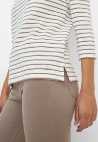 Gestreept langeroef top in wit en bruin, gladde stof. Beigekleurige broek met een aansluitende pasvorm en zijnaaddetails, hand rustend op de taille.