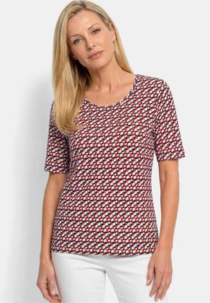 Femme aux longs cheveux blonds portant une chemise rouge, noire et blanche à manches courtes avec un motif géométrique, et un pantalon blanc, debout devant un fond uni.