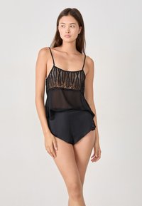 Le modèle pose dans un camisole en dentelle noire et soie avec un short assorti, mettant en valeur des détails complexes et une coupe décontractée, sur un fond clair.