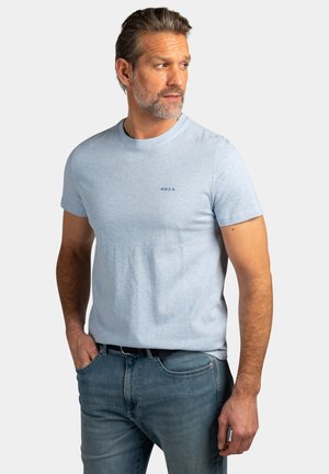 Man met kort grijs haar en baard, gekleed in een lichtblauw T-shirt met korte mouwen en een blauwe spijkerbroek, kijkt naar links met zijn handen in de zakken.