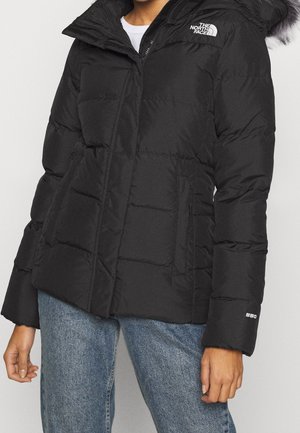 Chaqueta acolchada negra con capucha forrada de piel, bolsillos delanteros y el logo de The North Face, usada con vaqueros azules.