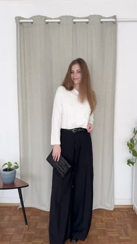 Λευκό ριμπάριστο τοπ μακρυά μανίκια, μαύρο παντελόνι καμπάνα με ψηλή μέση, κρατώντας μια μαύρη τσάντα φάκελο με υφή, στέκεται σε ξύλινο πάτωμα, μπέζ κουρτίνα.