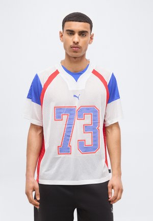Young man wearing white mesh sports jersey with blue and red accents and the number 73, standing against a plain background.