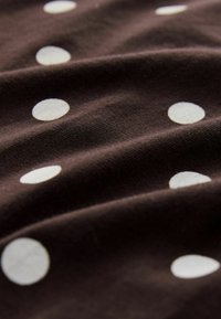 Tissu en coton de couleur marron foncé avec des pois blancs. La texture est lisse avec des plis et des rides visibles. Les pois sont uniformément espacés.