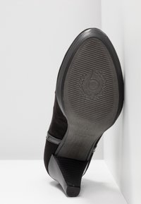 Chaussure noire à talon positionnée semelle vers le haut contre un mur blanc, montrant une semelle texturée avec un logo circulaire et un petit talon empilé.