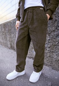 Hnědé manšestrové kalhoty s vertikálními pruhy, elastickým pasem, bočními kapsami, spárované s bílými teniskami na texturovaném povrchu.