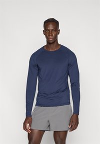 Αθλητική μπλούζα με μακρύ μανίκι σε ναυτικό μπλε ύφασμα με στρογγυλή λαιμόκοψη, με λεία υφή και εφαρμοστή γραμμή, συνδυασμένη με κοντό παντελόνι ανοιχτού γκρι χρώματος.