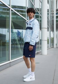 Giacca a vento leggera azzurro chiaro con un motivo e una striscia blu navy, abbinata a pantaloni corti blu navy e sneakers bianche. Design chiaro e leggero; vestibilità casual.