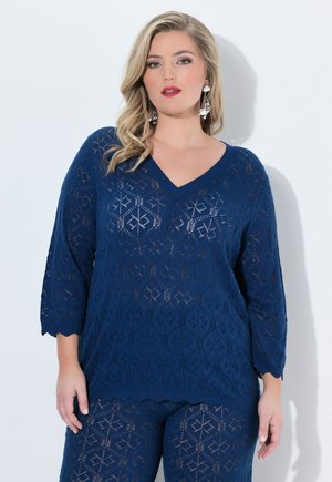 Frau trägt blau gemustertes Stricktop und passende Hose, mit welligem blondem Haar und großen silbernen Ohrringen, vor weißem Hintergrund.