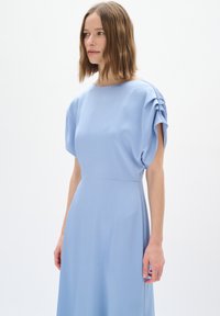 Abito azzurro chiaro con maniche corte e a sbuffo e scollatura rotonda semplice. Tessuto strutturato, vita aderente e gonna fluida.
