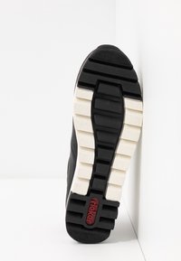 Semelle d'une basket noire et blanche avec des rainures profondes et le logo rouge "Rieker" au talon, positionnée verticalement sur un fond blanc.