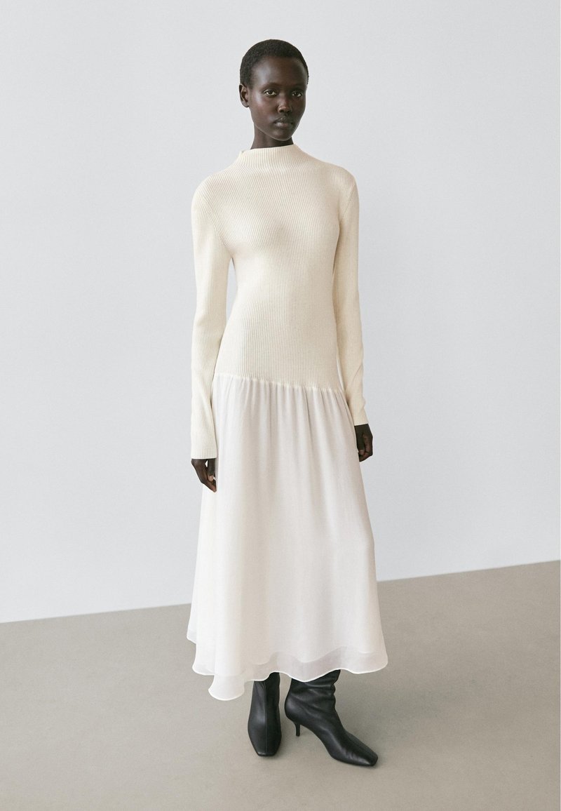 Maglione color avorio a coste con collo alto che si trasforma in una gonna fluente in chiffon bianco; abbinato a stivaletti neri.