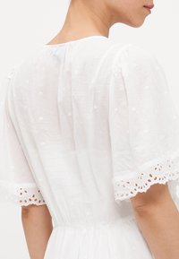 Abito bianco in cotone con scollo rotondo, maniche corte a sbuffo, dettagli floreali ricamati e pizzo lungo i bordi delle maniche.