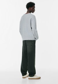 Nuori mies seisoo sivuttain, päällään vaaleanharmaa paita, löysät mustat farkut ja valkoiset lenkkarit, taustanaan yksinkertainen valkoinen tausta.