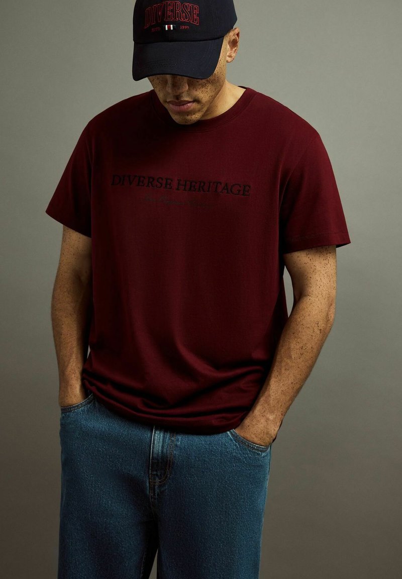 Bordowa koszulka z krótkim rękawem z napisem "DIVERSE HERITAGE" w czarnym kolorze, zestawiona z niebieskimi dżinsami oraz granatową czapką baseballową z czerwonymi akcentami.