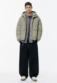 Nafukovacia bunda v svetlo olivovej zelenej farbe, s objemom, kapucňou a prednými vreckami. Nosená cez károvanú košeľu a čierne široké džínsy.