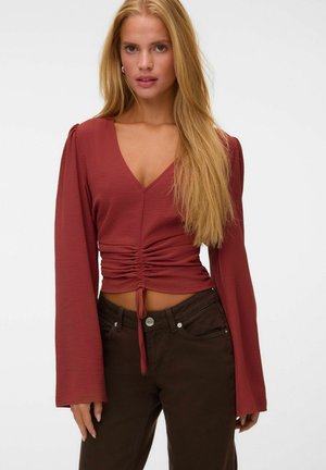 Femme aux longs cheveux blonds portant un chemisier froncé couleur rouille à manches longues et un pantalon marron foncé sur un fond uni.