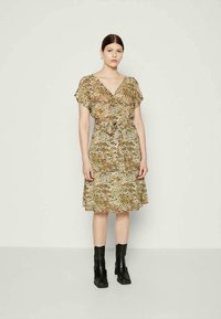 Vestido bege com padrão de leopardo, mangas curtas, decote em V, cintura amarrada e barra rodada. Usado com botas de tornozelo pretas. Tecido suave e leve.