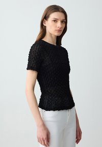 Top nero lavorato a maglia con un motivo testurizzato e maniche corte, caratterizzato da un colletto rotondo e una vita aderente, abbinato a pantaloni bianchi.