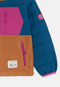 Pametna jakna s modrim zgornjim delom, rožnatimi detajli in rjavim flisom spodaj. Ima sprednji žep, vrvico in detajl norveške zastave.